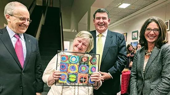 An artist holding a piece of calendar art surrounded by two men and one woman.