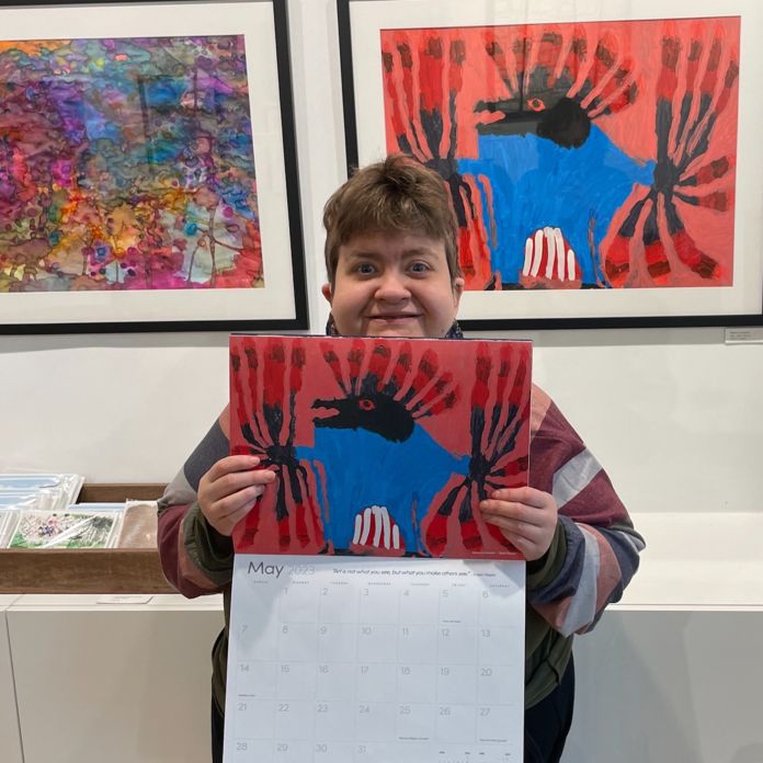 A female artist holding a 2023 calendar in front of her artwork of a blue figure with feathers on a red background.